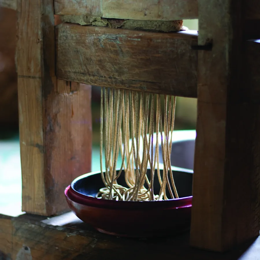 Buckwheat noodles being pressed through a wooden press into a bowl