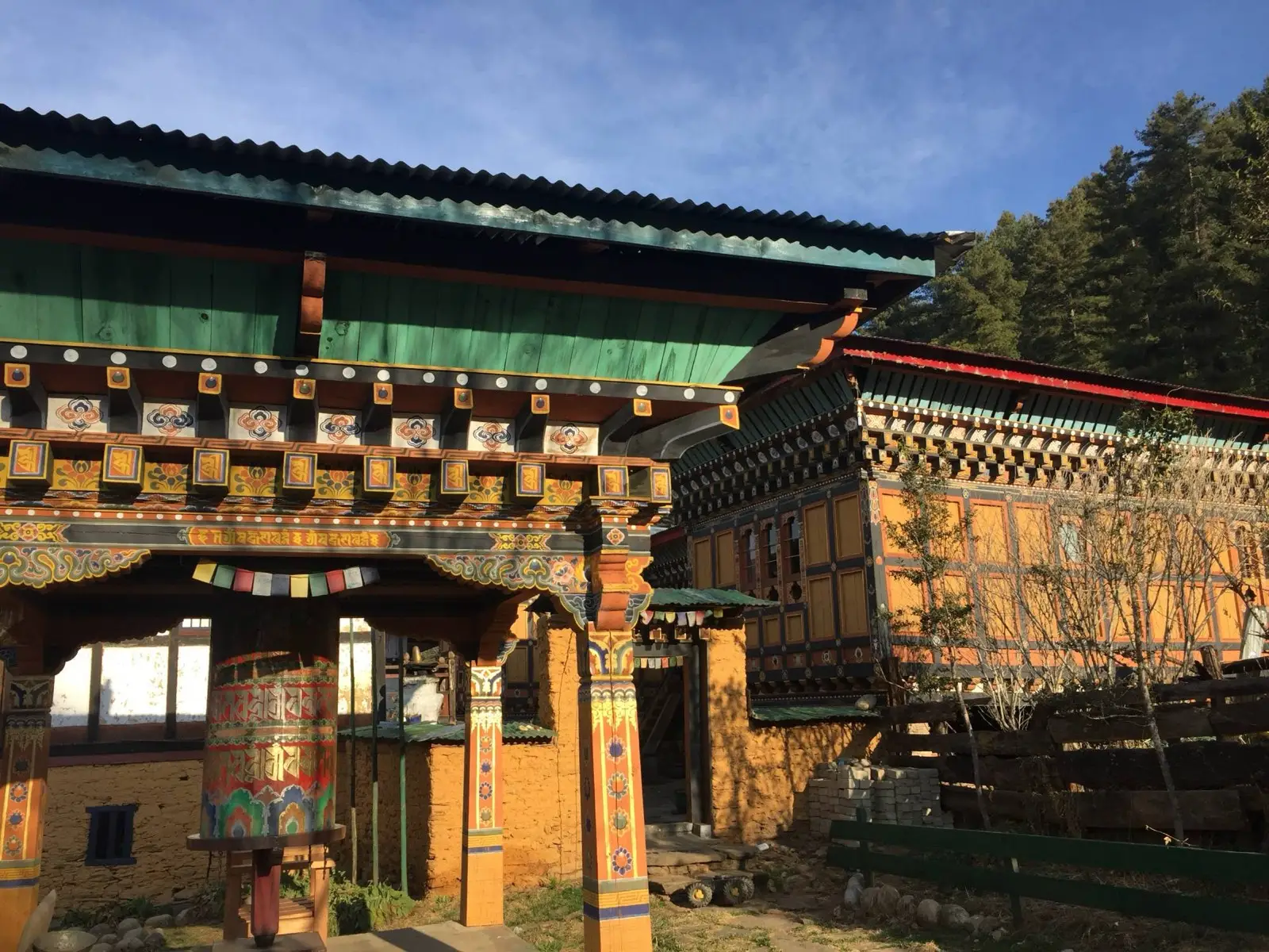 Painted temple courtyard with a large prayer wheel and a traditional house in morning sun